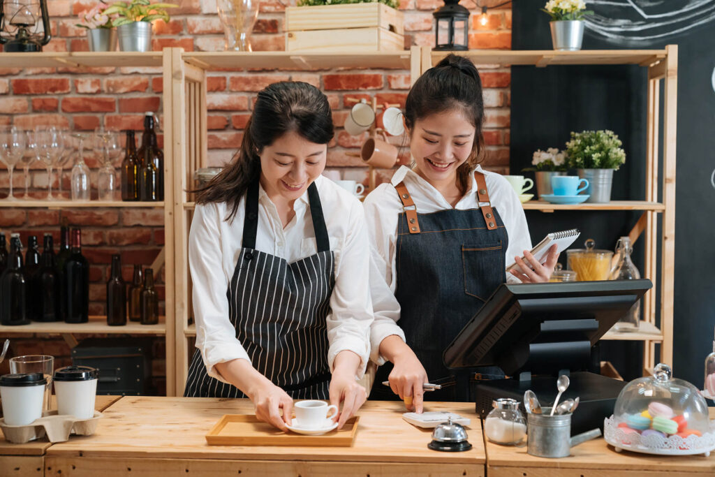 Two women side by side wearing aprons operating a coffee shop.