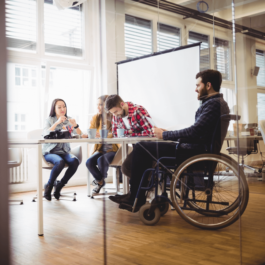 Group of four people, one of them in a wheelchair, sitting around a table with coffee cups and a blank projection screen