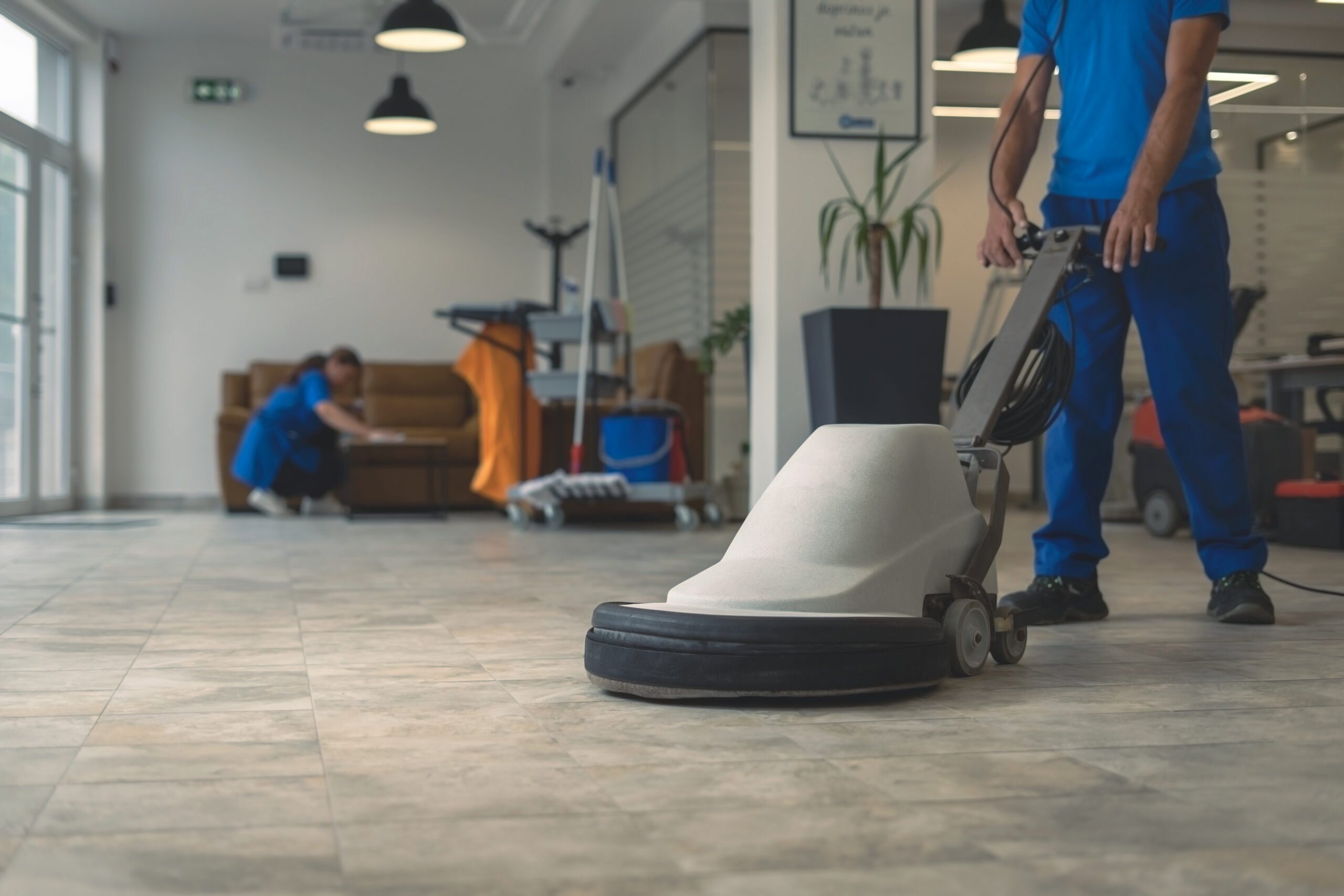 Worker polishing hard floor with high-speed polishing machine while other cleaner cleans the table in the background