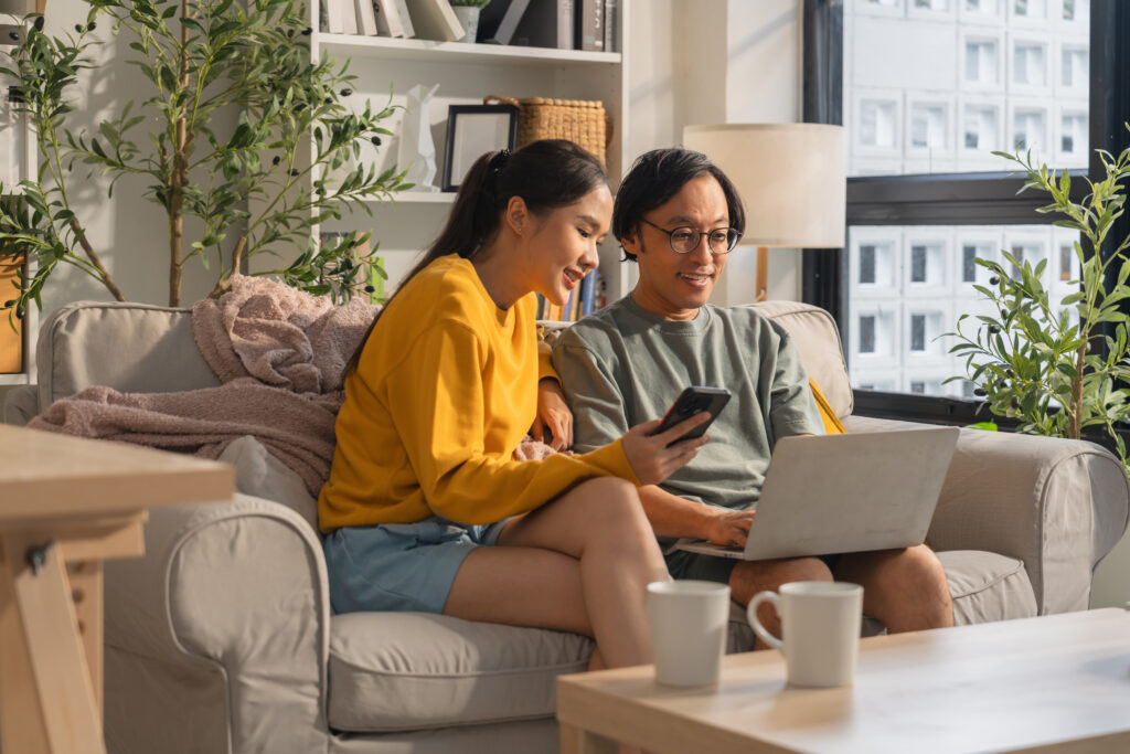 Una pareja asiática casada pasa tiempo de calidad juntos en un sofá en la sala de estar de su casa, usando alegremente una computadora portátil y un teléfono inteligente para mirar Quaint Oak Bank tasas de certificados de depósito